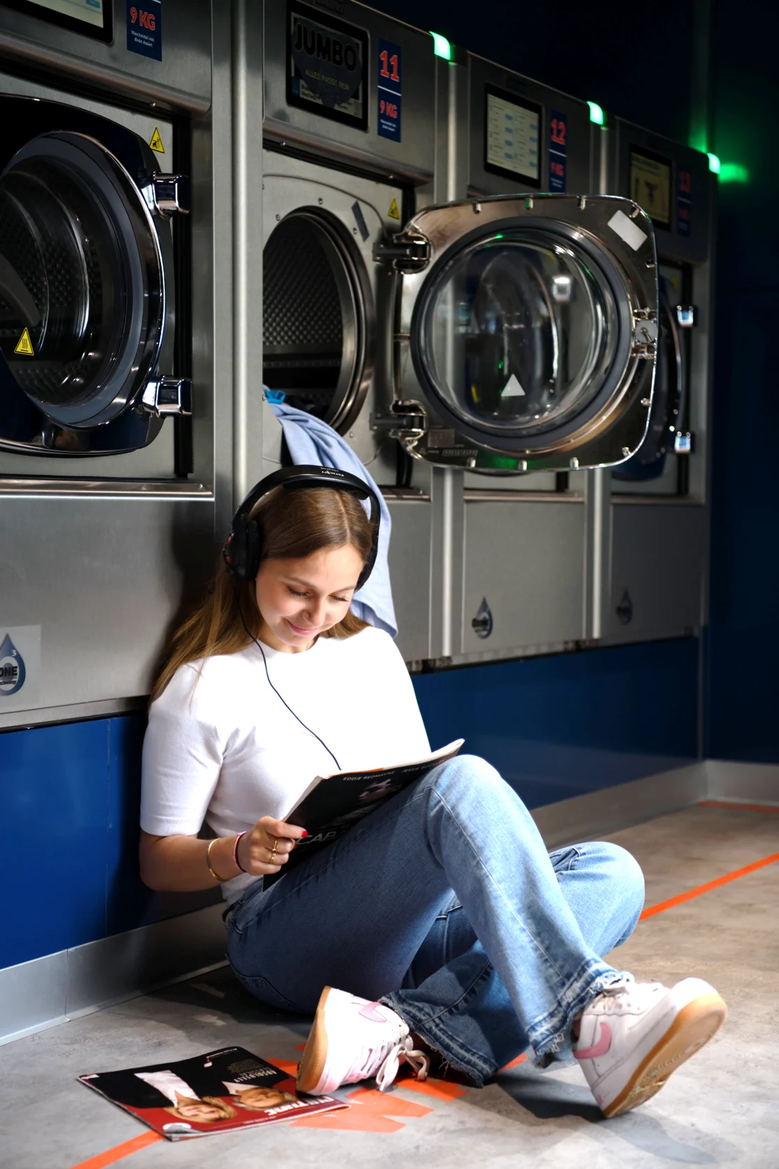 Young woman relaxing and reading while doing laundry in a Girbau self-service laundromat with modern automatic washing machines.