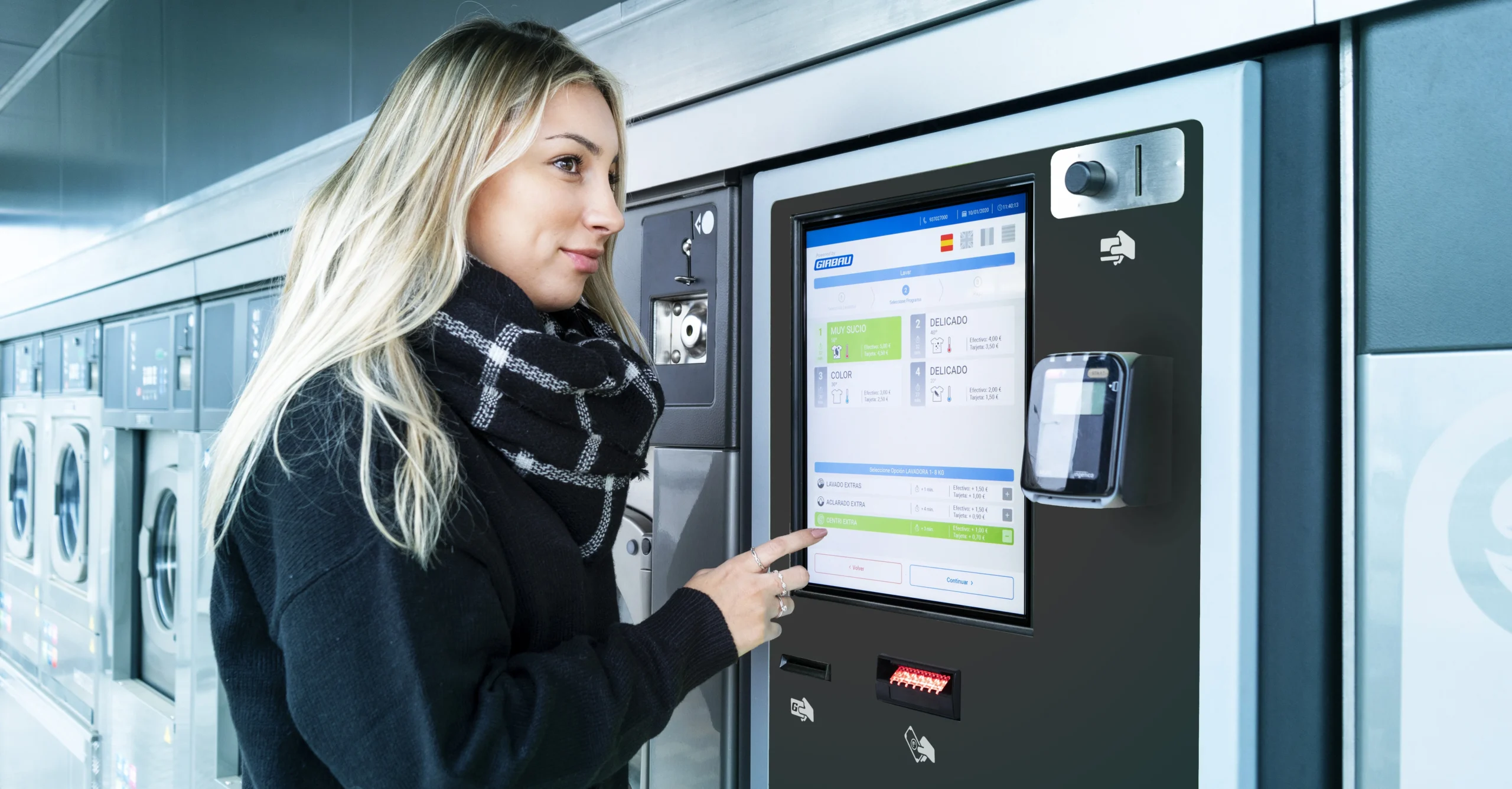 Customer using a Girbau self-service laundry payment kiosk with touch-screen interface inside an automatic laundromat.