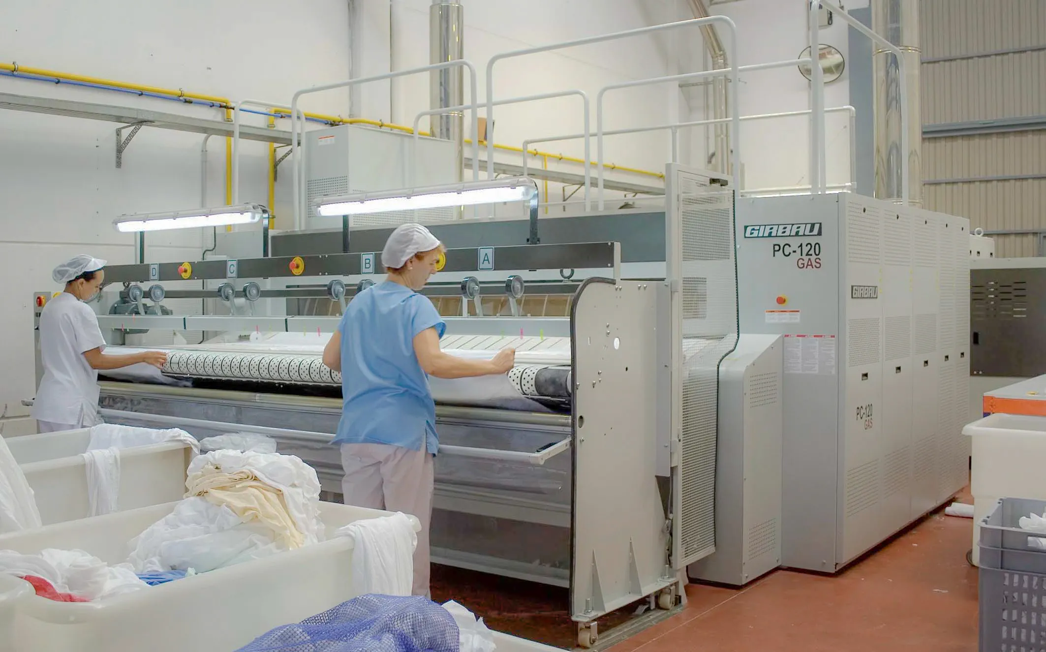 Industrial laundry operators feeding linens into a Girbau flatwork ironer, representing large-scale linen finishing processes.
