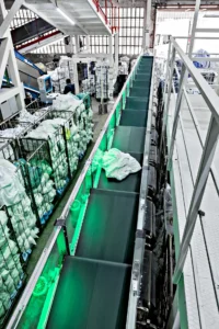 Vista superior del sistema de clasificación de ropa Sortech con cintas transportadoras moviendo distintos tipos de textiles, mostrando una operación de alta capacidad en un espacio de lavandería compacto.