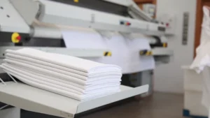 Ropa plana apilada junto a la planchadora industrial compacta Compact Pro, representando el cumplimiento de normativas, la fabricación certificada y la garantía oficial Girbau.