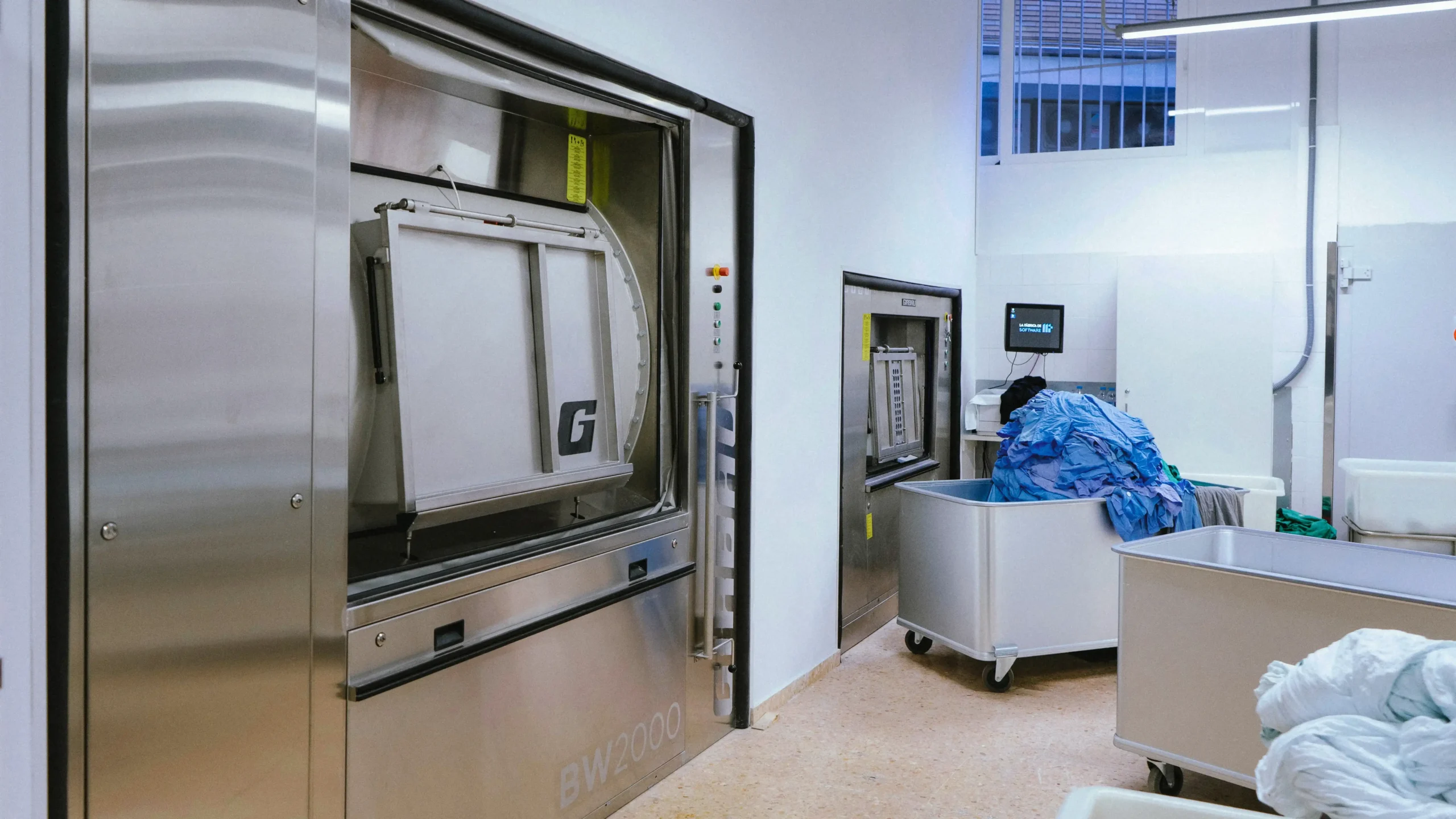 Blanchisserie hospitalière avec laveuses à barrière sanitaire et chariots de linge médical, illustrant les solutions Girbau pour un lavage sûr et hygiénique.