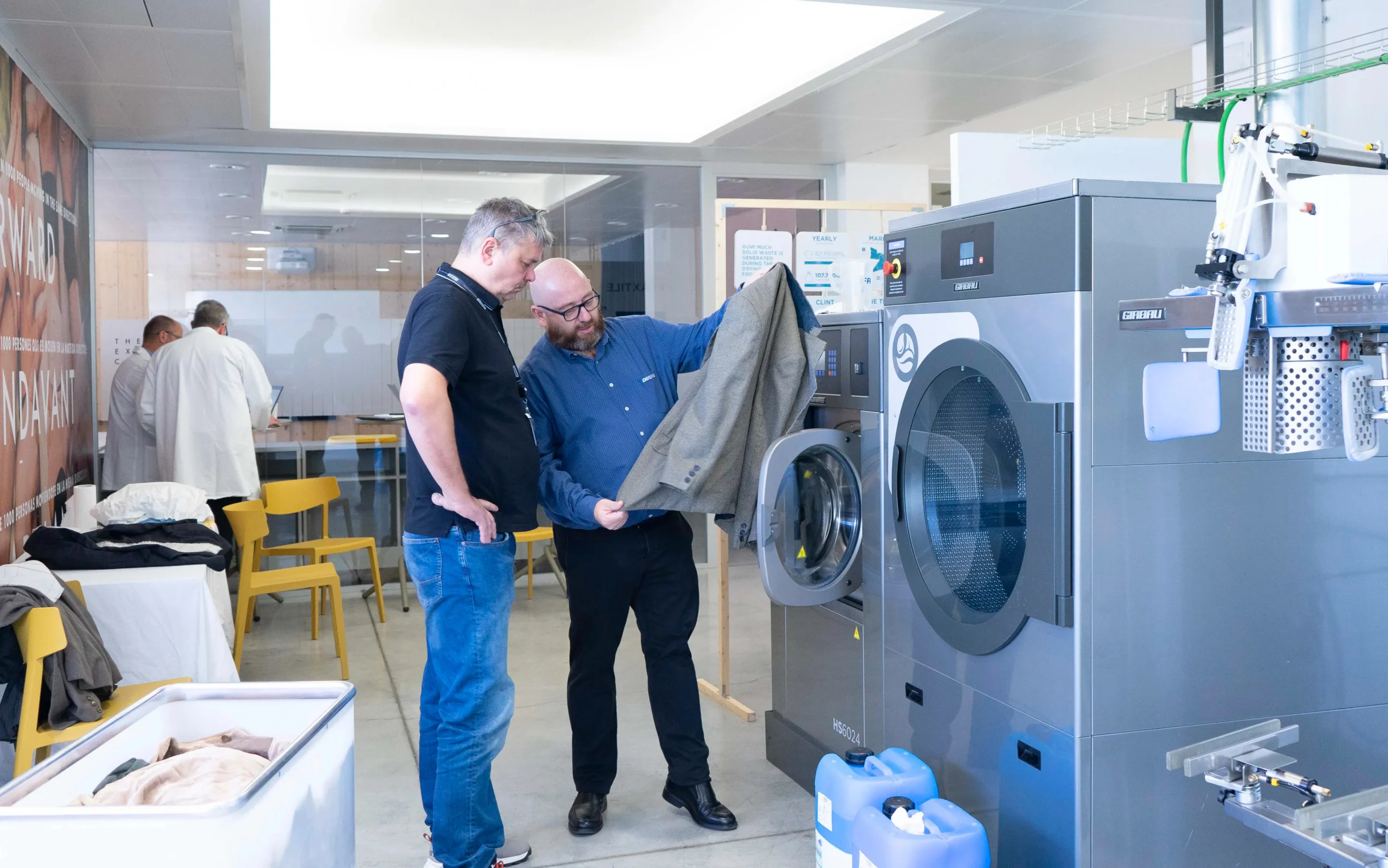 Techniciens inspectant un vêtement à côté d’équipements de nettoyage à l’eau Girbau, représentant l’évaluation et le nettoyage professionnel des textiles.