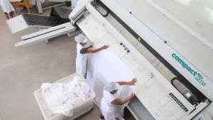 Operarios utilizando la planchadora industrial compacta Compact Pro para procesar prendas planas de gran tamaño, destacando su capacidad, adaptación a espacios reducidos y bajo consumo energético.