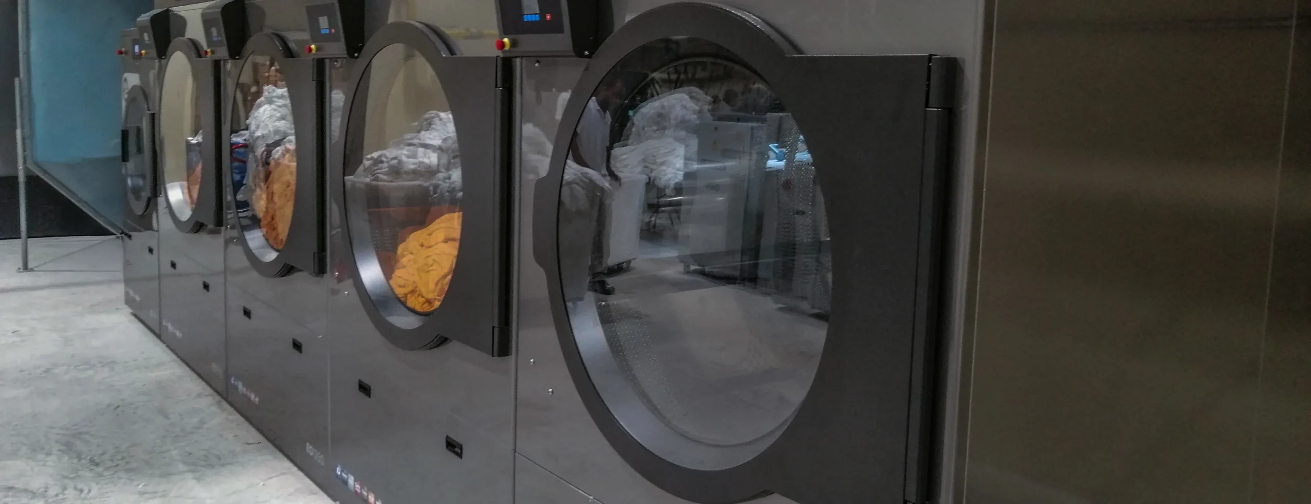 Row of high-capacity commercial dryers operating in a professional laundry, showing large front-loading doors and efficient textile drying systems for industrial use.